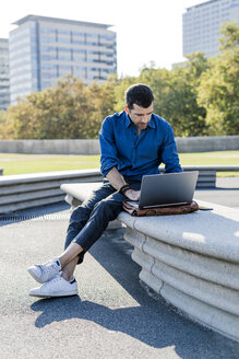 Geschäftsmann sitzt auf einer Bank im Freien und arbeitet an einem Laptop - GIOF05707
