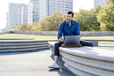 Smiling businessman sitting on bench outdoors working on laptop - GIOF05708