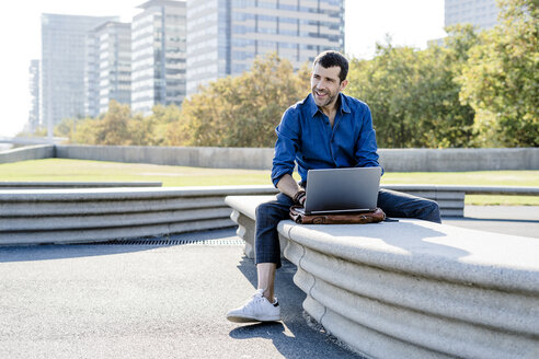 Lächelnder Geschäftsmann sitzt auf einer Bank im Freien und arbeitet an einem Laptop - GIOF05708