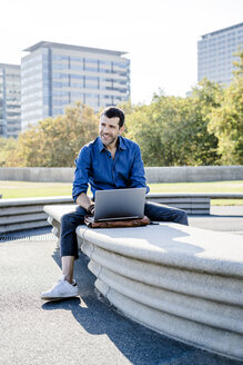 Porträt eines lächelnden Geschäftsmannes, der auf einer Bank im Freien sitzt und an einem Laptop arbeitet - GIOF05709