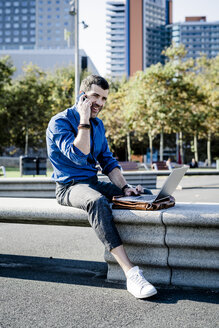 Lächelnder Geschäftsmann am Telefon sitzt auf Bank im Freien mit Laptop - GIOF05710