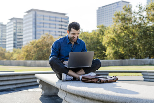 Lächelnder Geschäftsmann sitzt auf einer Bank im Freien und arbeitet an einem Laptop - GIOF05711