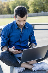 Businessman sitting on bench outdoors using smartphone and laptop - GIOF05713