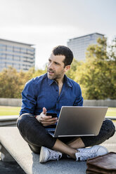 Businessman sitting on bench with smartphone and laptop looking at distance - GIOF05714