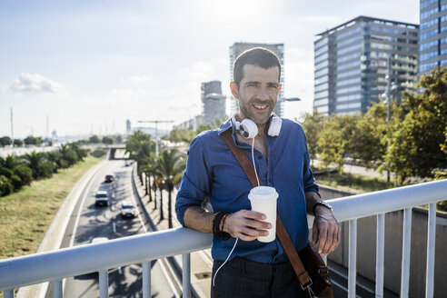 Porträt eines lächelnden Mannes mit Kaffee zum Mitnehmen und Kopfhörern, der an einem Brückengeländer lehnt - GIOF05729