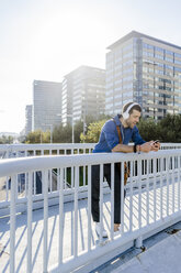 Singing man leaning on railing of footbridge listening music with headphones and smartphone - GIOF05733