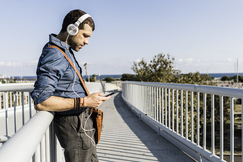 Mann steht auf einer Fußgängerbrücke und hört Musik mit Kopfhörern und Smartphone - GIOF05736