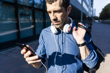 Porträt eines Mannes mit Kopfhörern und Tasche, der auf sein Mobiltelefon schaut - GIOF05739