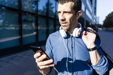 Porträt eines Mannes mit Kopfhörern, Tasche und Mobiltelefon, der in die Ferne schaut, lizenzfreies Stockfoto