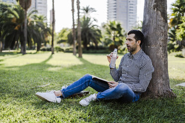 Man on a meadow in city park leaning against tree drinking water - GIOF05747