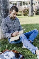 Man on a meadow leaning against tree trunk taking notes - GIOF05751