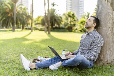 Man with laptop leaning against tree trunk on meadow in city park relaxing - GIOF05755