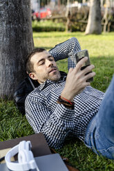 Portrait of man lying on meadow in city park looking at cell phone - GIOF05758