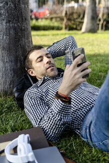 Porträt eines Mannes, der auf einer Wiese im Stadtpark liegt und auf sein Mobiltelefon schaut - GIOF05758