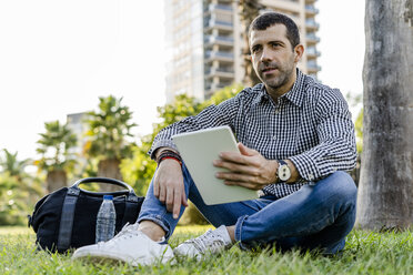 Porträt eines Mannes mit digitalem Tablet, der auf einer Wiese im Stadtpark sitzt und denkt - GIOF05759