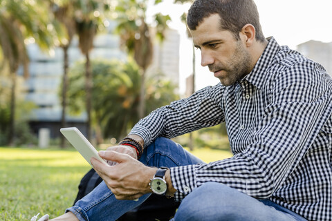 Mann sitzt auf einer Wiese im Stadtpark und schaut auf ein digitales Tablet, lizenzfreies Stockfoto