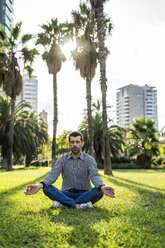 Man sitting on meadow in city park doing yoga exercise - GIOF05764