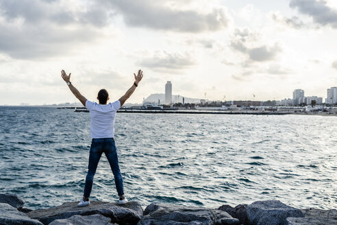 Spain, Barcelona, back view of man with raised arms standing on rock in front of the sea - GIOF05770