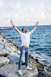Mann mit erhobenen Armen und geschlossenen Augen vor dem Meer stehend - GIOF05772