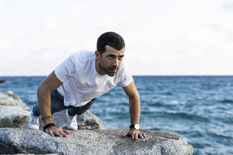 Mann macht Liegestütze auf einem Felsen vor dem Meer, lizenzfreies Stockfoto