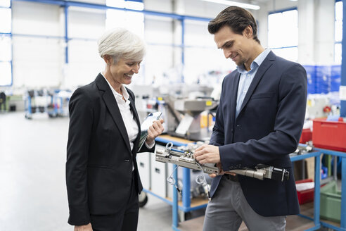 Ein lächelnder Geschäftsmann und eine ältere Geschäftsfrau begutachten ein Werkstück in einer Fabrik - DIGF05701