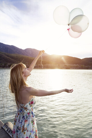 Junge Frau im Sommerkleid mit Blumenmuster steht auf einem Steg und betrachtet Luftballons, lizenzfreies Stockfoto