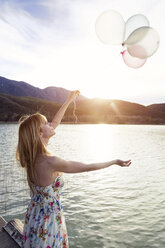 Young woman wearing summer dress with floral design standing on jetty looking at balloons - JSRF00110