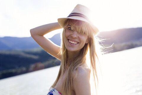 Porträt einer lächelnden jungen Frau vor einem See mit Sommerhut in der Abenddämmerung, lizenzfreies Stockfoto