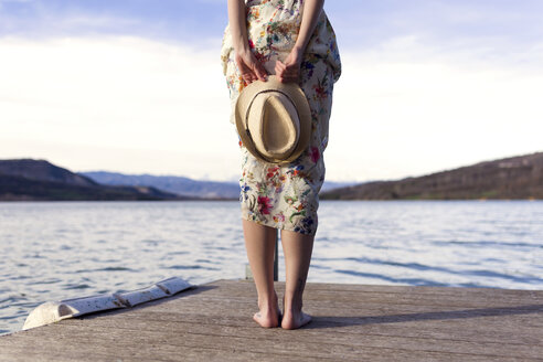 Rückenansicht einer barfüßigen jungen Frau, die auf einem Steg steht und einen Sommerhut hält, Teilansicht - JSRF00115