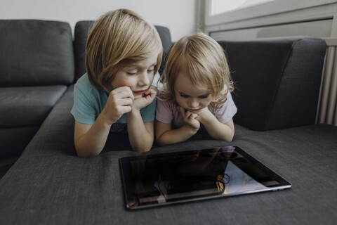 Bruder und kleine Schwester liegen zu Hause auf der Couch und benutzen ein digitales Tablet, lizenzfreies Stockfoto