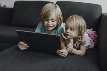 Portrait of brother and little sister lying on the couch at home using digital tablet - JLOF00301