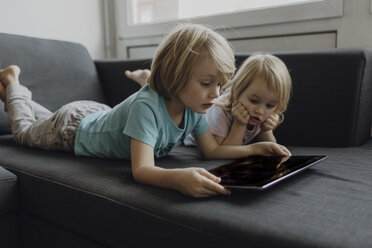 Brother and little sister lying on the couch at home using digital tablet - JLOF00302