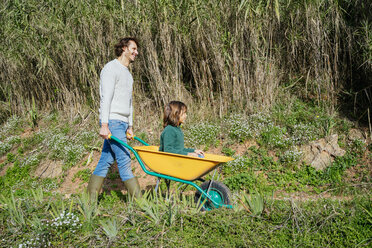 Father walking on a dirt track, pushing wheelbarrow, with his son sitting in it - GEMF02704