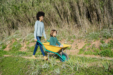 Father walking on a dirt track, pushing wheelbarrow, with his son sitting in it - GEMF02705