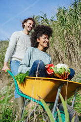 Woman sitting in wheelbarrow, holding fresh vegetables, man pushing her - GEMF02708