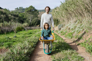 Father walking on a dirt track, pushing wheelbarrow, with his son sitting in it - GEMF02780