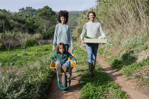 Familie geht auf einem Feldweg, schiebt Schubkarre, trägt Kiste mit Gemüse, lizenzfreies Stockfoto