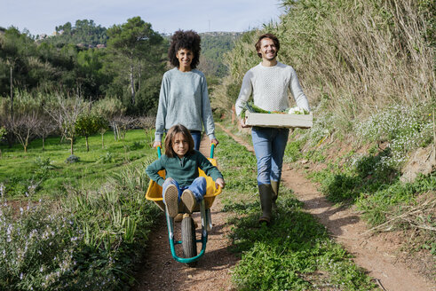 Family walking on a dirt track, pushing wheelbarrow, carrying crate with vegetables - GEMF02783