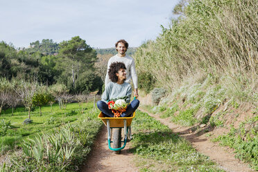 Woman sitting in wheelbarrow, holding fresh vegetables, man pushing her - GEMF02784