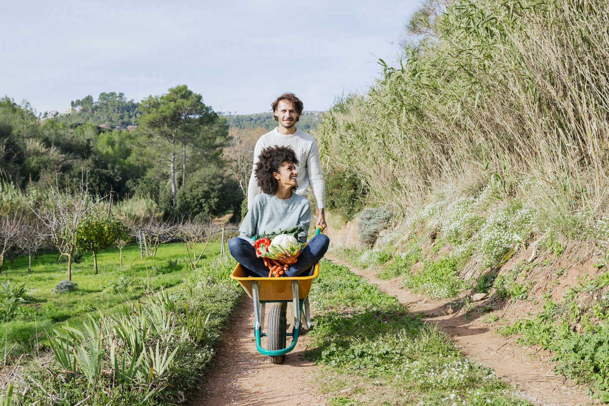 Woman sitting in wheelbarrow holding fresh vegetables man