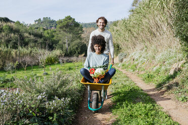 Woman sitting in wheelbarrow, holding fresh vegetables, man pushing her - GEMF02785