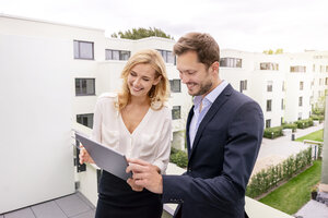 Immobilienmakler, der mit einem Kunden auf einem Balkon steht und auf ein digitales Tablet schaut - PESF01462