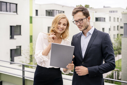 Real estate agent standing on a balcony with customer, looking at digital tablet - PESF01465