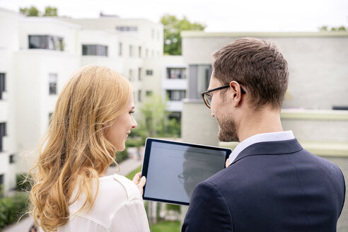 Immobilienmakler, der mit einem Kunden auf einem Balkon steht und auf ein digitales Tablet schaut - PESF01467