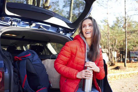 Glückliche Frau, die während einer Autoreise mit einer Thermoskanne im Kofferraum sitzt, lizenzfreies Stockfoto