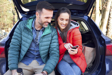 Couple using smartphone while sitting in car trunk during road trip - BSZF00903