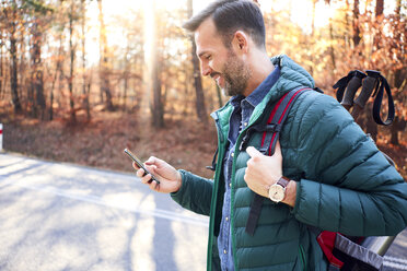 Lächelnder Mann, der auf einer Straße im Wald während einer Rucksacktour sein Smartphone überprüft - BSZF00911