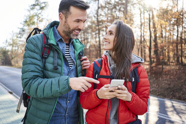 Glückliches Paar, das auf einer Straße im Wald während einer Rucksacktour sein Smartphone überprüft - BSZF00912