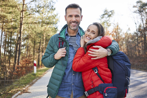 Porträt eines glücklichen Paares, das sich auf einer Straße im Wald während einer Rucksacktour umarmt, lizenzfreies Stockfoto