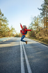 Cheerful woman jumping on an empty road during backpacking trip - BSZF00917